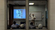 Through two windows, a teacher in a white shirt stands in a classroom in front of a whiteboard and beside a screen with a slide on it. Four students wearing blue shirts and two with white caps on face the teacher with their backs to the windows.