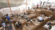 Several people dig and sift through brown dirt, surrounded by black buckets and tools.