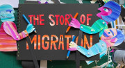 Three colorful figures made of paper hold colored pencils and stand around a black book called “The Story of Migration.” A drafting table covered in scraps of paper fills the background.