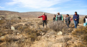 A person in a red shirt points out a spot to a group of people standing among dry grasses and rocks.