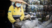 A person wearing a straw hat, blue face mask, yellow shirt, and gloves sorts plastic waste in a bin in front of stacked bundles of multicolored plastic.