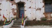 A white temple wall with partially exposed brick is covered with holes. Two red, green, and yellow dragons perch on either side of stairs leading to a blue door.