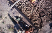 In an aerial view, two people kneel and dig at opposite sides of a square plot of dirt, each beside buckets, pickaxes, and trowels. Between them is a large pile of differently shaped bones.